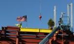 Closeup of yellow beam with evergreen & American flag and worker on top of red steel framework of new 2-story building