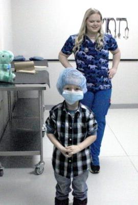 孩子戴着外科口罩和帽子，里面有毛绒玩具，背景是穿着手术服的大学生